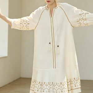 May include: Off-white dress with embroidered details. The dress features a V-neckline, long sleeves, and a knee-length hemline. The embroidery is in a contrasting tan color, with geometric patterns on the sleeves and hem. The dress has a relaxed fit.