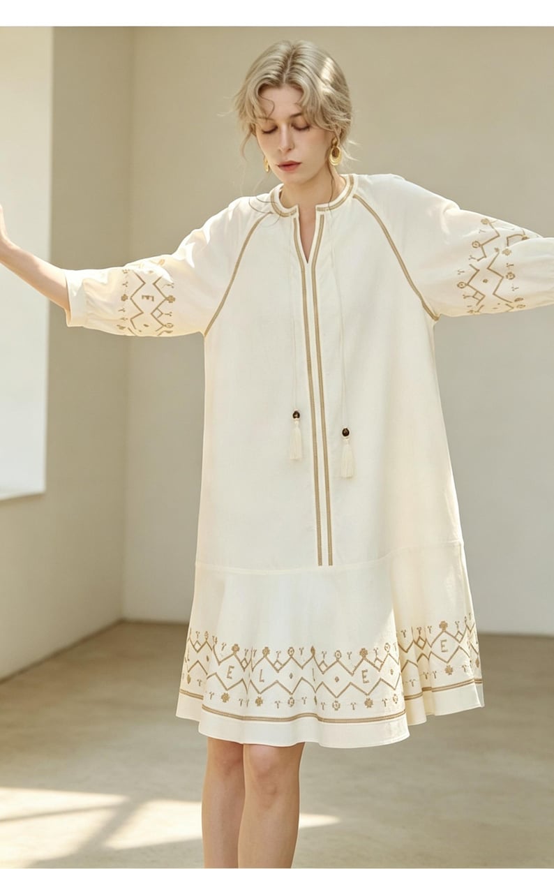 May include: Off-white dress with embroidered details. The dress features a V-neckline, long sleeves, and a knee-length hemline. The embroidery is in a contrasting tan color, with geometric patterns on the sleeves and hem. The dress has a relaxed fit.