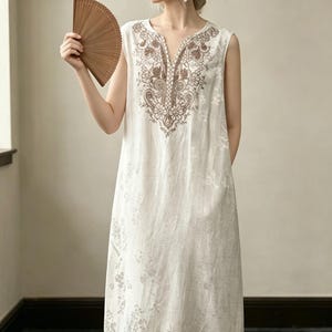 May include: An off-white sleeveless dress with a V-neck and brown embroidery. The dress has a floral pattern and a brown border. A wooden fan is held in the hand. The dress is approximately 130 cm in length.