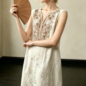 May include: A sleeveless beige dress with brown floral embroidery around the neckline and bodice. The dress has a V-neck and a subtle floral pattern on the skirt. A brown fan is held in the left hand.