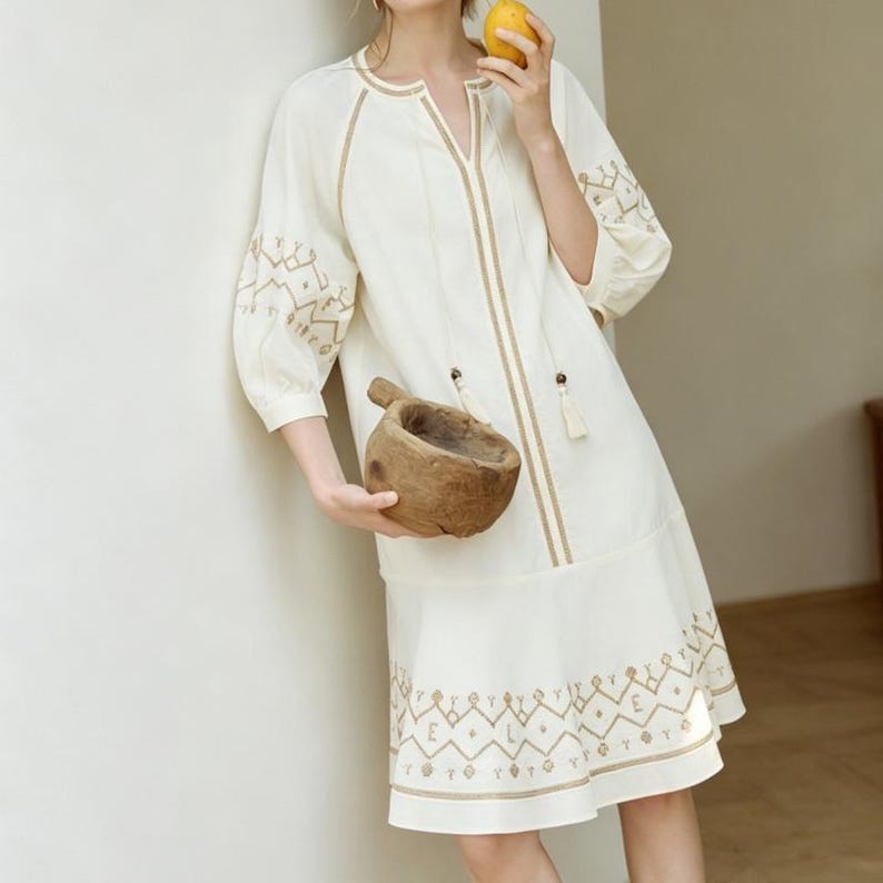 May include: Cream-colored dress with embroidered details. The dress features a V-neck, three-quarter sleeves, and a tiered skirt. The embroidery is in a contrasting tan color, with geometric patterns on the sleeves and hem. A wooden bowl is held in the image.