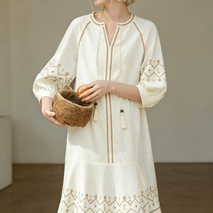 May include: Cream-colored dress with intricate brown embroidery. The dress features a V-neck, three-quarter sleeves, and a flared hem. The embroidery includes geometric and floral patterns. The dress is accessorized with tassels and is suitable for casual wear.