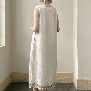 May include: A cream-coloured, sleeveless dress featuring a delicate floral pattern. The dress has a simple, elegant design with a slightly flared silhouette. The hemline is decorated with a decorative border. Made from lightweight fabric, ideal for warmer weather.