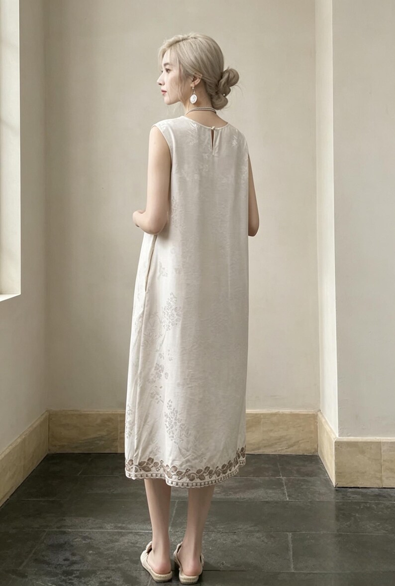 May include: A cream-coloured, sleeveless dress featuring a delicate floral pattern. The dress has a simple, elegant design with a slightly flared silhouette. The hemline is decorated with a decorative border. Made from lightweight fabric, ideal for warmer weather.