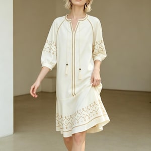 May include: Off-white dress with gold embroidery. The dress features a V-neck, three-quarter sleeves, and a flared hemline. Gold embroidery accents the neckline, sleeves, and hem. The dress has a relaxed fit and is made of a lightweight fabric.