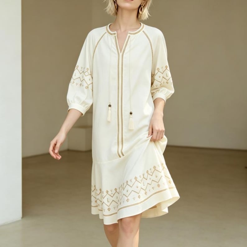 May include: Off-white dress with gold embroidery. The dress features a V-neck, three-quarter sleeves, and a flared hemline. Gold embroidery accents the neckline, sleeves, and hem. The dress has a relaxed fit and is made of a lightweight fabric.