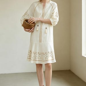 May include: Cream-colored embroidered dress with gold accents. The dress features a V-neck, tassel details, and geometric embroidery on the sleeves and hem. The person is holding a small woven basket.