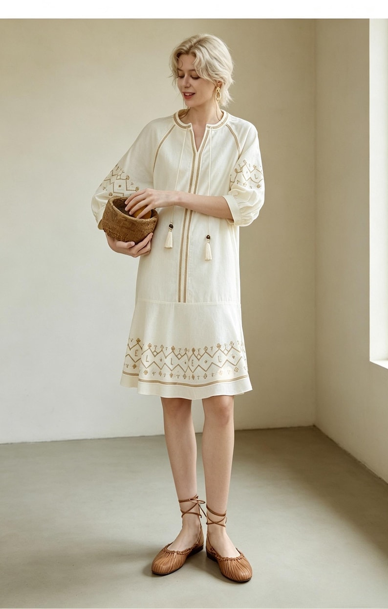 May include: Cream-colored embroidered dress with gold accents. The dress features a V-neck, tassel details, and geometric embroidery on the sleeves and hem. The person is holding a small woven basket.