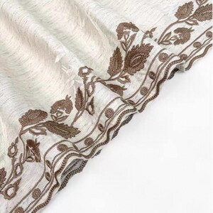 May include: Off-white fabric with a detailed brown floral embroidery border. The embroidery features leaves and flower-like designs. The fabric has a textured appearance, and the border is intricately stitched.