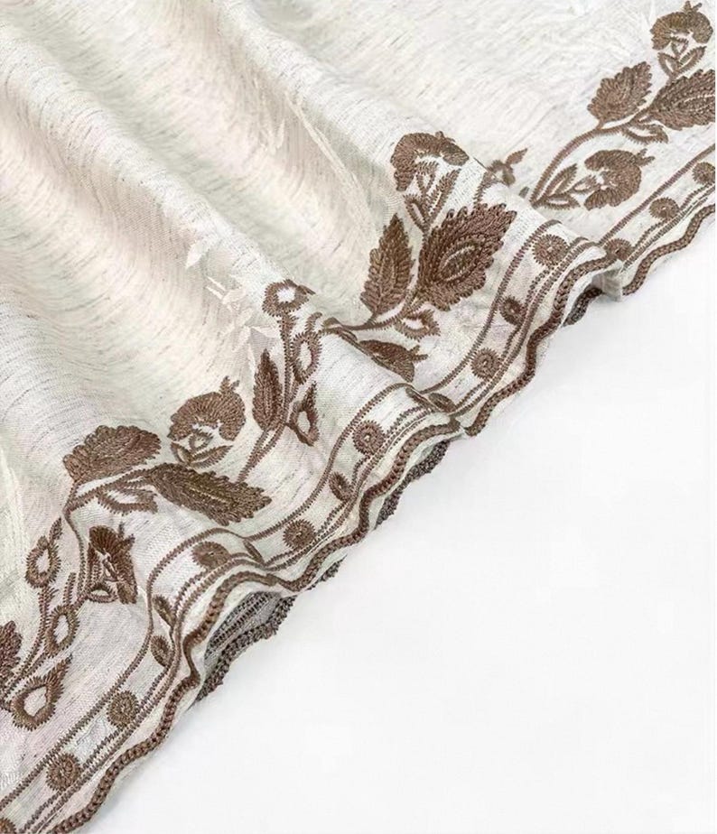 May include: Off-white fabric with a detailed brown floral embroidery border. The embroidery features leaves and flower-like designs. The fabric has a textured appearance, and the border is intricately stitched.