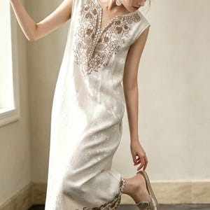 May include: An off-white sleeveless dress with brown embroidery around the neckline and hem. The dress has a V-neck and a subtle floral pattern. The model is wearing beige slip-on shoes.