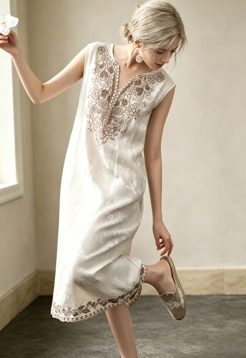May include: An off-white sleeveless dress with brown embroidery around the neckline and hem. The dress has a V-neck and a subtle floral pattern. The model is wearing beige slip-on shoes.