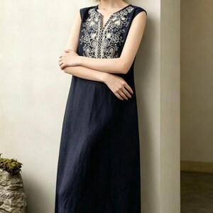 May include: A navy blue sleeveless dress with white embroidered detailing around the neckline and chest. The dress has a V-neck and a flowing, A-line shape. The embroidery showcases floral and geometric designs.