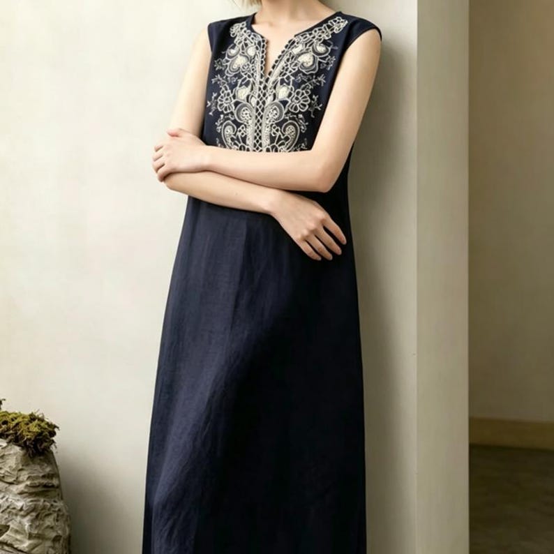 May include: A navy blue sleeveless dress with white embroidered detailing around the neckline and chest. The dress has a V-neck and a flowing, A-line shape. The embroidery showcases floral and geometric designs.
