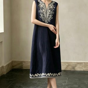 May include: A navy blue sleeveless dress with white embroidery. The dress has a V-neck and floral detailing on the neckline and hem. The knee-length dress is made of a lightweight fabric. The model is wearing white shoes.