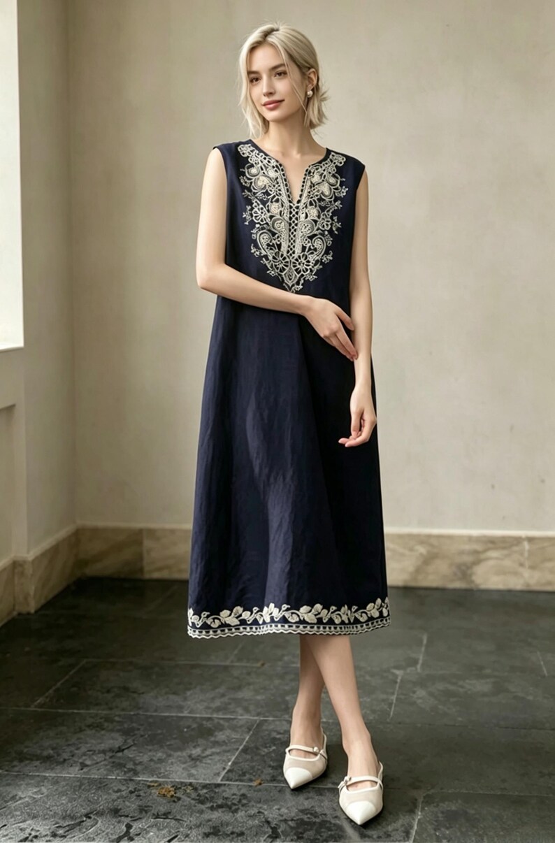 May include: A navy blue sleeveless dress with white embroidery. The dress has a V-neck and floral detailing on the neckline and hem. The knee-length dress is made of a lightweight fabric. The model is wearing white shoes.