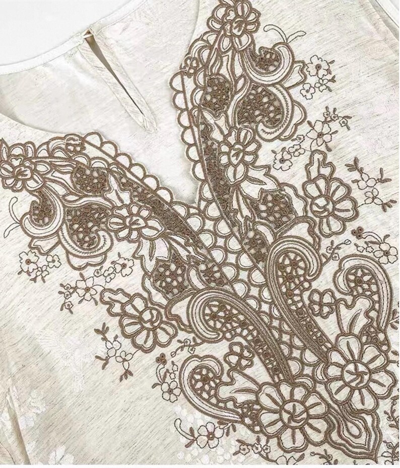 May include: Close-up of a light-coloured top with a V-neckline. The neckline and upper chest area are adorned with intricate brown floral lace embroidery. The fabric has a subtle textured appearance.