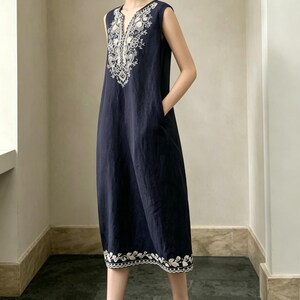 May include: A navy blue sleeveless dress with white embroidered detailing around the neckline and hem. The dress has a V-neck and pockets. The model is wearing white and beige slip-on shoes.
