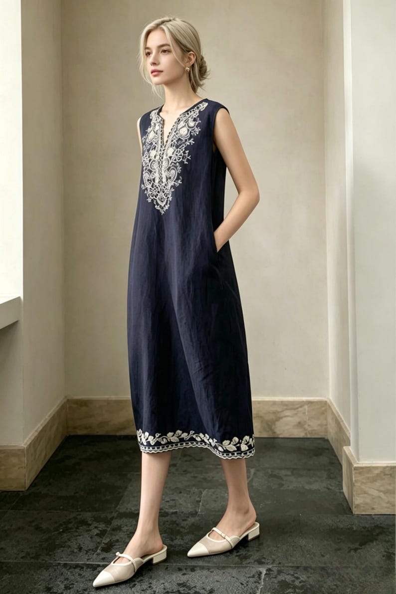 May include: A navy blue sleeveless dress with white embroidered detailing around the neckline and hem. The dress has a V-neck and pockets. The model is wearing white and beige slip-on shoes.
