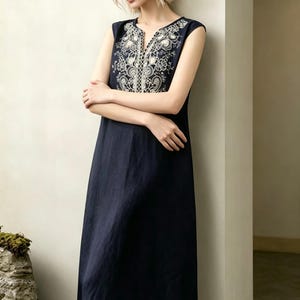 May include: A navy blue sleeveless dress featuring white floral embroidery on the neckline and hem. The dress has a V-neck and a relaxed fit. The model is leaning against a wall. The dress is made of a soft, flowing fabric.