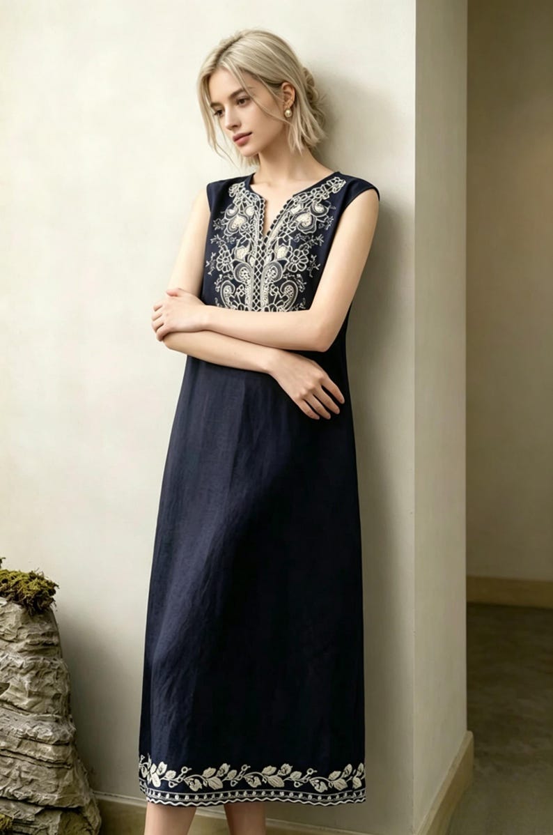 May include: A navy blue sleeveless dress featuring white floral embroidery on the neckline and hem. The dress has a V-neck and a relaxed fit. The model is leaning against a wall. The dress is made of a soft, flowing fabric.