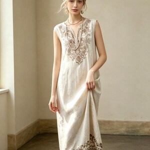 May include: An off-white sleeveless dress featuring detailed brown embroidery on the neckline and hem. The dress has a loose fit and a delicate floral design. The model is wearing a necklace and earrings.