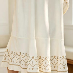 May include: Cream-colored dress with intricate gold embroidery along the hem and sleeves. The dress features a flared skirt and a detailed geometric pattern, creating a bohemian aesthetic. The sleeves also have gold embroidery.