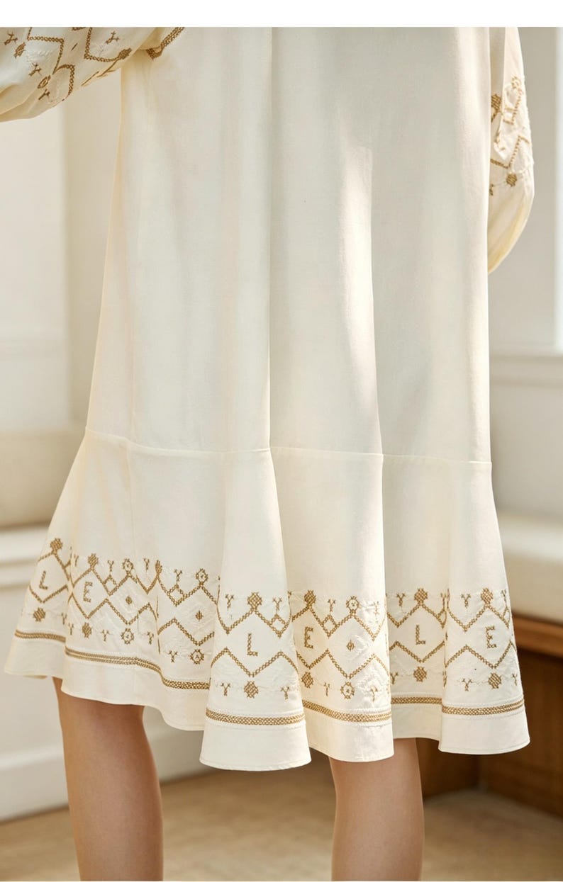May include: Cream-colored dress with intricate gold embroidery along the hem and sleeves. The dress features a flared skirt and a detailed geometric pattern, creating a bohemian aesthetic. The sleeves also have gold embroidery.