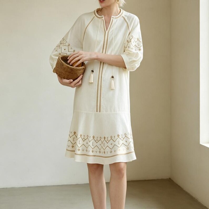May include: Cream-colored dress with embroidered details. The dress features a V-neck, long sleeves, and a flared hem. The embroidery is in a contrasting tan color, with geometric patterns on the sleeves and hem. Two tassels hang from the neckline.