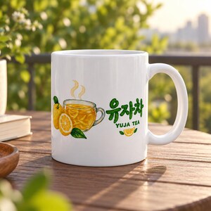 以下が含まれることがあります： 黄色いお茶が入ったガラスのカップ、丸ごとのユズ、スライスしたレモン、緑の葉が描かれた白いセラミック製のマグカップ。緑色で「유자차」と「YUJA TEA」という文字が印刷されており、その横には小さなレモンのスライスのグラフィックが添えられています。
