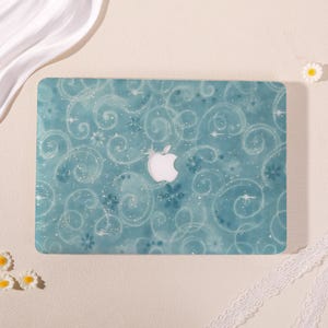 Peut inclure: Une coque rigide pour ordinateur portable de couleur sarcelle avec un motif de tourbillons blancs, des accents en forme d'étoiles et des fleurs. La coque présente une découpe centrale révélant le logo Apple argenté pour protéger l'appareil.