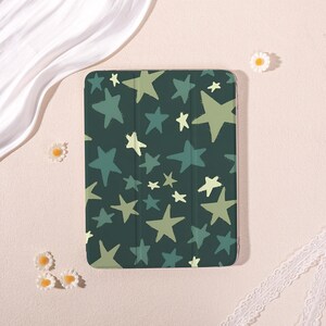May include: A rectangular tablet case featuring a dark forest green background patterned with scattered five-pointed stars in varying shades of light sage green, pale yellow, and muted teal, with a smooth finish and visible vertical fold lines.