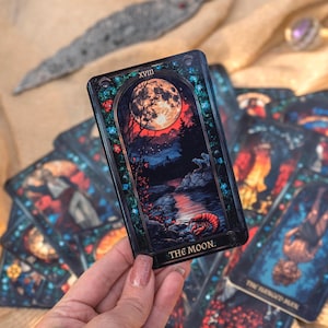 May include: A tarot card from a deck, featuring a full moon over a dark landscape with a river and trees. The card is labeled "THE MOON" and has a decorative border. Other cards are visible in the background.