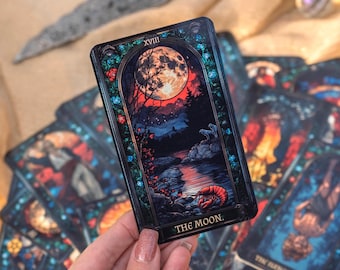 May include: A tarot card from a deck, featuring a full moon over a dark landscape with a river and trees. The card is labeled "THE MOON" and has a decorative border. Other cards are visible in the background.