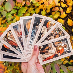 May include: A hand holding a fan of tarot cards with detailed illustrations. The cards feature images of angels, figures, and celestial bodies. The cards have a cream-colored border and are set against a backdrop of autumn leaves.