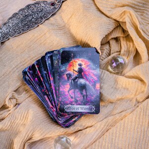 May include: A deck of tarot cards with a fantasy illustration of a figure on horseback, labeled "Page of Wands." The cards are fanned out on a gold-colored cloth. A decorative silver incense holder and a crystal ball are also present.