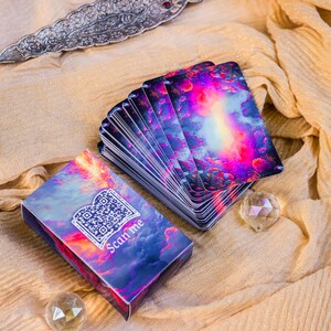 May include: A deck of tarot cards with a celestial design, featuring vibrant pink, purple, and blue hues. The accompanying box displays a QR code and the text "Scan me." Two crystal spheres and a decorative silver blade are also present.