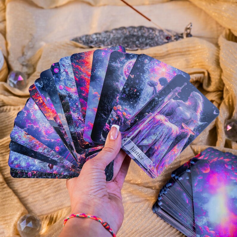 May include: A hand holding a fanned deck of tarot cards. The cards feature celestial and fantasy artwork in shades of purple, blue, and red. The card at the front reads "Justice". A second deck of cards is visible in the background.