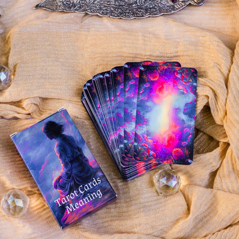 May include: A deck of tarot cards with a box featuring a person in a flowing robe against a purple and blue sky. The cards have a vibrant, colorful design with pink, purple, and blue hues. The box reads "Tarot Cards Meaning".