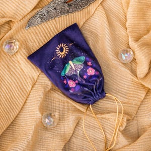 May include: A dark purple velvet drawstring bag featuring a moth and floral design with a sun and moon. The bag has a gold drawstring and is set on a gold, textured fabric. Decorative crystals and a silver blade are also present.