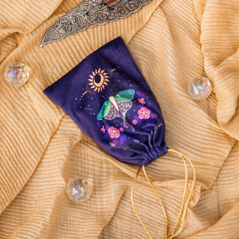 May include: A dark purple velvet drawstring bag featuring a moth and floral design with a sun and moon. The bag has a gold drawstring and is set on a gold, textured fabric. Decorative crystals and a silver blade are also present.