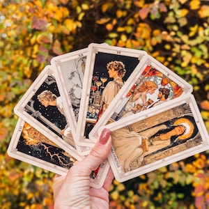 May include: A hand holding a fan of tarot cards with illustrations in a vintage style. The cards feature images of people, lightning, and architectural elements. The background is blurred with autumn foliage.