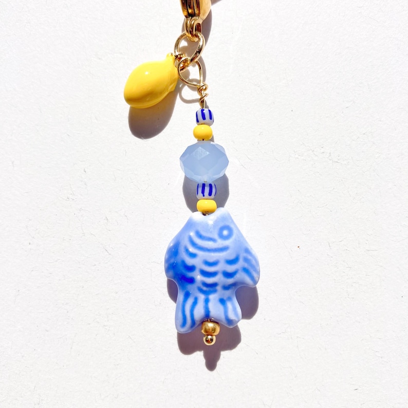 May include: A whimsical charm featuring a blue ceramic fish pendant with white details, accented by yellow and blue beads. A yellow teardrop-shaped charm hangs above. The charm is attached to a gold-colored clasp.
