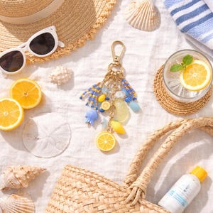 May include: A beach-themed still life featuring a straw hat, sunglasses, and a woven tote bag. A decorative keychain with lemon charms and blue beads is displayed with seashells, lemon slices, and a refreshing drink.