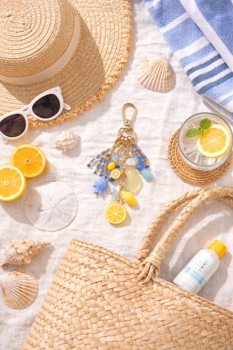 May include: A beach-themed still life featuring a straw hat, sunglasses, and a woven tote bag. A decorative keychain with lemon charms and blue beads is displayed with seashells, lemon slices, and a refreshing drink.