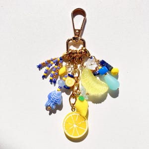May include: A gold-colored keychain with a lobster clasp, adorned with charms. Charms include yellow lemon slices, a blue fish, yellow and blue beads, and a yellow lemon wedge. Additional charms include blue and yellow beads and a light blue teardrop.