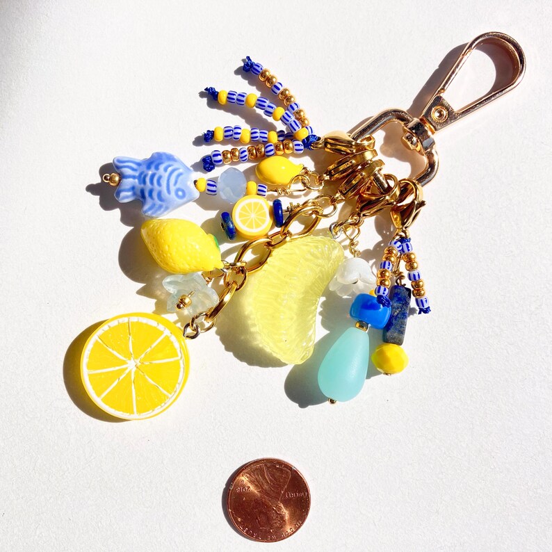 May include: A gold-colored keychain with charms featuring a lemon slice, a lemon, a lemon wedge, and a blue fish. The keychain also includes blue, yellow, and clear beads, and a gold-colored clasp.