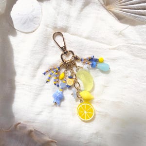 May include: A gold-colored keychain with a lobster clasp, adorned with lemon-themed charms. The charms include a lemon slice, a lemon, and a lemon slice, along with blue and yellow beads. The keychain is a fun accessory.
