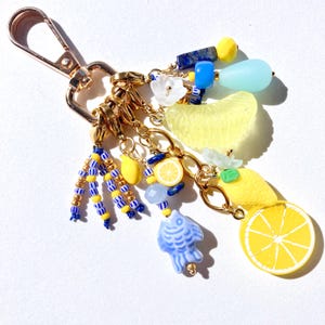 May include: A gold-toned keychain with various charms, including yellow lemon slices, blue fish, and beaded accents. The charms are connected by gold chains and a lobster clasp.