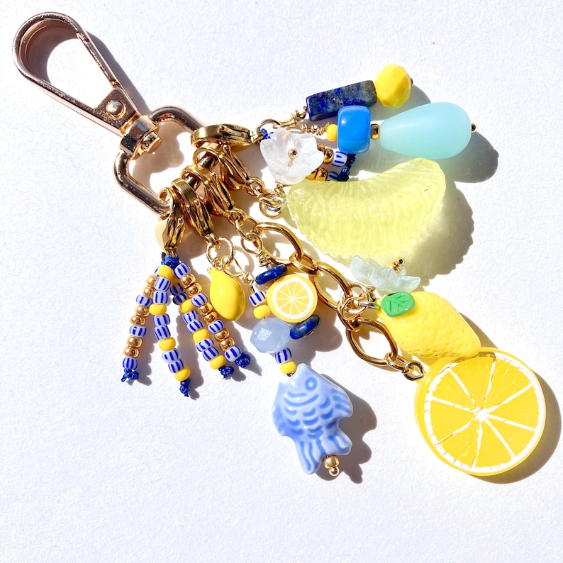 May include: A gold-toned keychain with various charms, including yellow lemon slices, blue fish, and beaded accents. The charms are connected by gold chains and a lobster clasp.